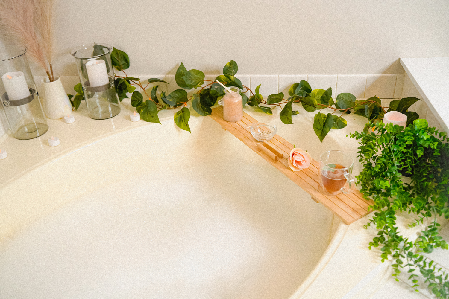 Relaxing bubble bath with candles and green vines around bath. Bath has wooden plank with tea, rose, and bath soak container
